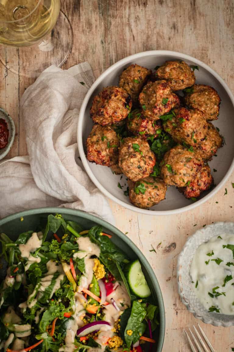 A bowl of meatballs sits on a timber table with a side salad a glass of white wine.