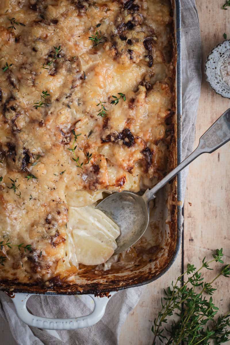 A cream enamel cast iron roasting pan full of cheesy potato bake. A spoonful has been eaten out of the corner.