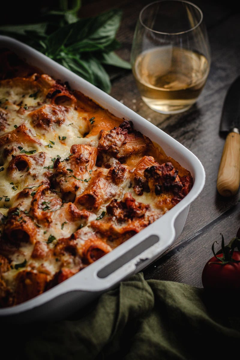 A large white enamel baking dish with a baked bolognese pasta bake in it. A glass of wine, a knife and some basil sit beside.